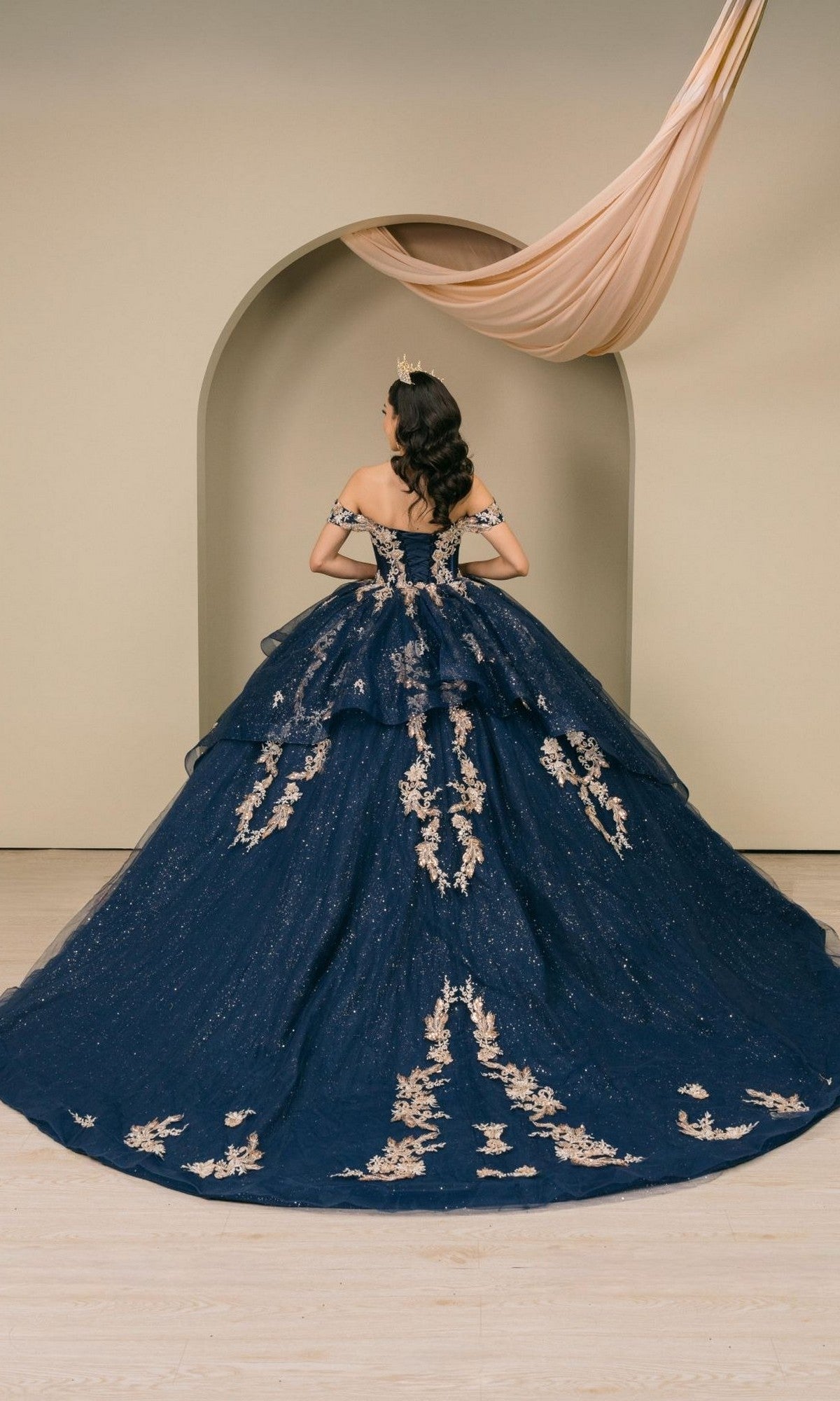 A woman in the Quinceanera Dress 8390JQ by Cinderella Couture, a navy ball gown adorned with gold embroidery, stands facing an arched alcove, her hair styled in loose waves and wearing a tiara.